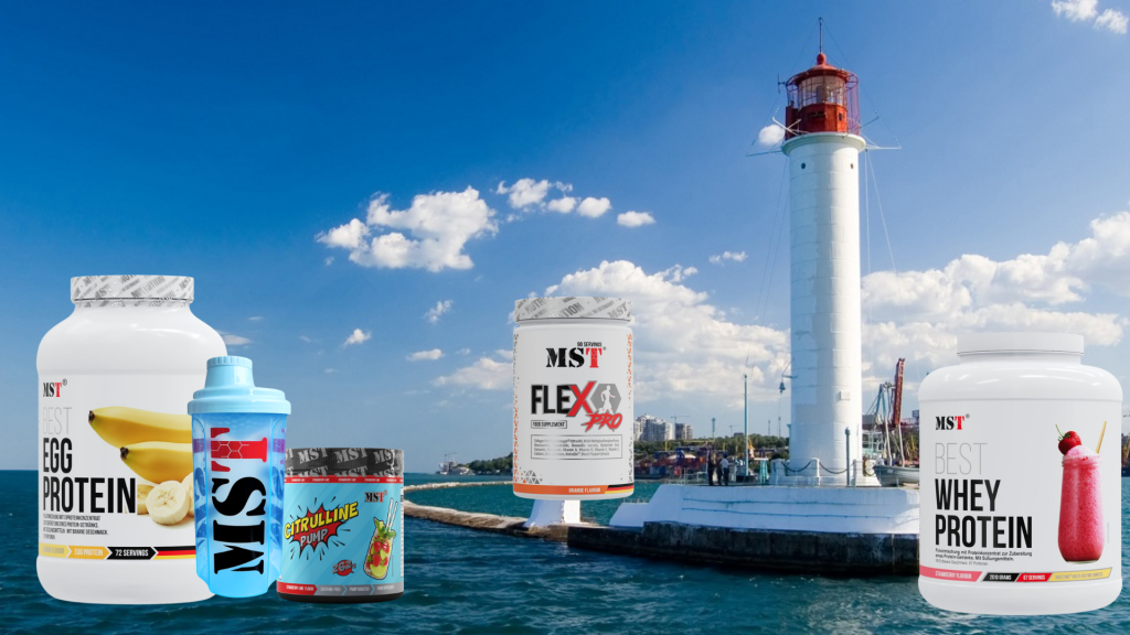 Рекламний колаж продукції бренду MST Nutrition на тлі морського пейзажу з білим маяком. На передньому плані зліва направо розміщено п'ять товарів: велика банка «Best Egg Protein» зі смаком банана, прозорий блакитний шейкер з логотипом MST, банка «Citrulline Pump», банка комплексу для суглобів «Flex Pro» та велика банка «Best Whey Protein» зі смаком полуниці. Продукти розташовані на тлі синього моря та кам'яного молу, що веде до високого біло-червоного маяка під ясним блакитним небом із білими хмарами.